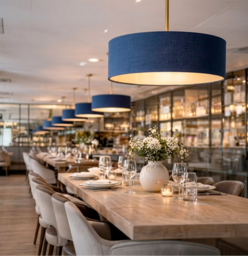 Large blue drum ceiling lampshades hanging above dining tables in a contemporary restaurant interior with warm ambient lighting.